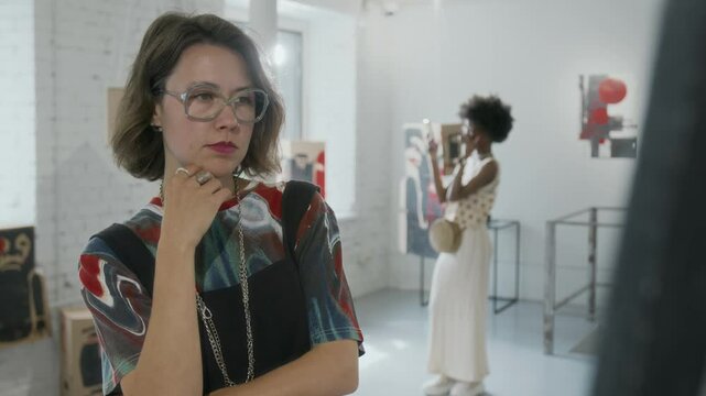 Medium shot of fashionably dressed young Caucasian woman in glasses, bright top, black bustier viewing conceptual painting at modern art exhibition, diverse visitors browsing artworks in background