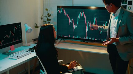 Male financial advisor gestures while explaining stock market fluctuations to a female client in a formal office setting at night, fostering an engaging discussion about investment strategies