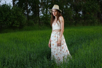 Beautiful girl in a hat and long dress dancing at sunset. Fashion portrait of young European model in green summer field.