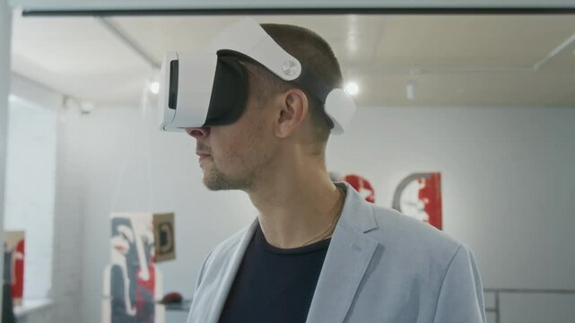 Medium closeup of young male visitor wearing VR headset looking at paintings at modern art exhibition in museum using virtual reality mobile application