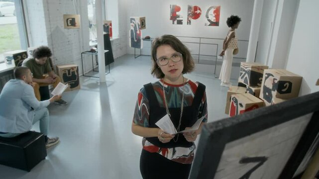 Medium full high-angle shot of young Caucasian female art critic standing in front of painting at modern art gallery, studying it, male visitors resting on benches, black woman viewing installation