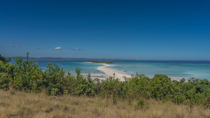 A sandy winding spit stretches towards a tropical island. Tiny silhouettes of people on the beach. Boats in the aquamarine ocean. In the foreground there is green vegetation - grass, bushes. 