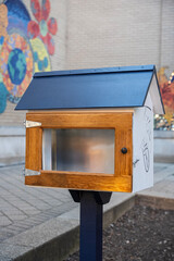 Wooden book exchange box with a metal roof set against a colorful mural, encouraging community sharing and literacy in an urban environment.