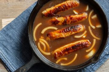 British pork sausages with onion gravy in a cast iron frying pan. On a wooden cutting board with a blue tea towel