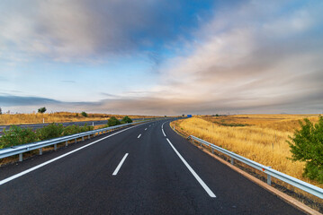 Fototapeta premium Empty highway between yellow grain fields and a dramatic sky at dawn, curved highway.