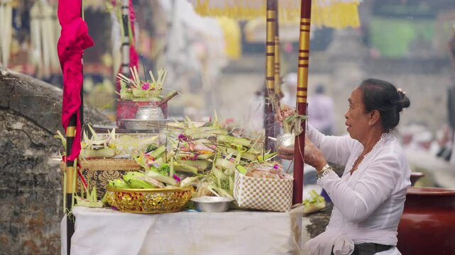 Woman sudra priest or pemangku in white traditional dress sprinkling holy water over offerings during kuningan ceremony in bali temple