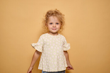 Charming toddler girl with wavy hair wearing beautiful shirt smiling with expressing pure innocence and happiness standing isolated over beige background