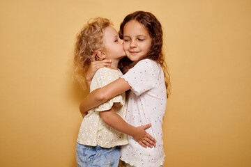 Curly blonde toddler embracing her brown haired sister with open hands both smiling wide and filled with happiness sharing joyful moment of love and support isolated on beige background