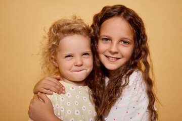 Blonde infant sharing joyful embrace with her brown-haired older sister while looking at the camera indoors displaying friendly and sociable spirit of childhood isolated over beige background