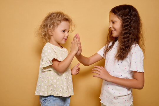 Wavy haired cheerful toddler clapping hands with her playful sister during active game of pat-a-cake both smiling carefree while cuddling together isolated on beige background
