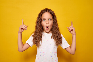 Little girl with wavy brown hair standing indoors against bright yellow background looking surprised and pointing up with excitement while displaying her amazement in advertising area