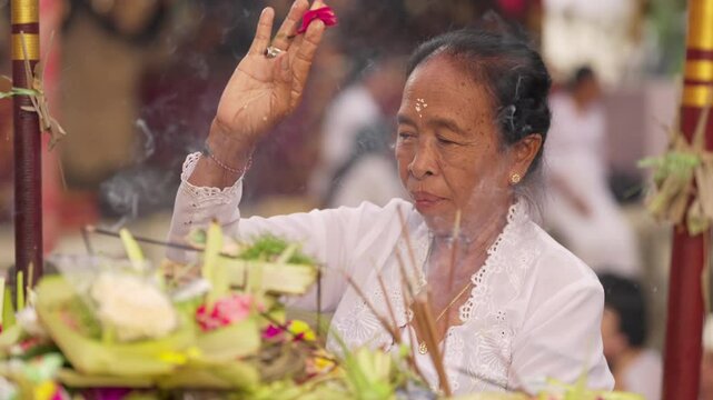 Balinese woman priest or pemangku performing traditional ritual at bali temple during kuningan religious celebration, offerings and burning incense