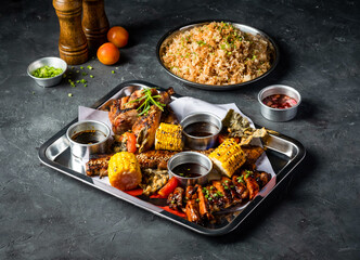 Filipino Feast with Grilled Meat, Garlic Fried Rice, grill corn cob, Peri Peri Chicken, Jerk Chicken, Beef Belly Brisket, Tomato and Sauces served tray isolated grey background side view of platter