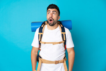 Young mountaineer man over isolated blue background looking up and with surprised expression