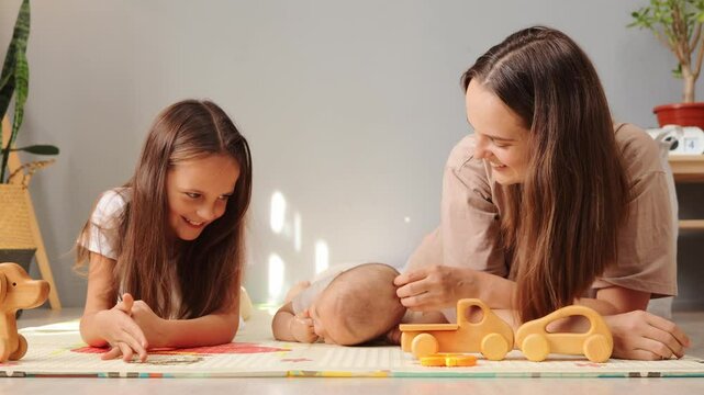 Pretty mother lying on the floor with her adorable newborn son and older daughter playing together with toys and enjoying moments of bonding indoors in living room