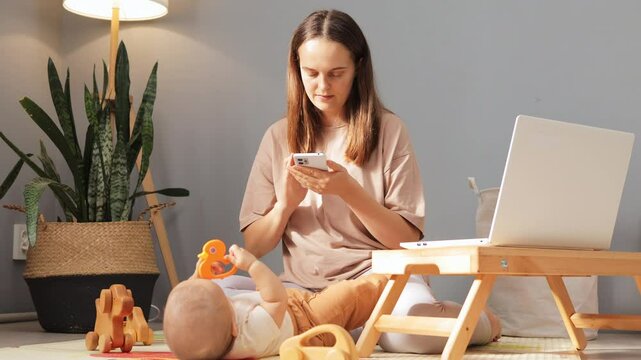 Busy mother using mobile phone while babysitting her daughter or son and working at home woman sitting on floor in living room engaging in online communication online browsing or internet shopping