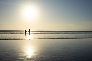 Fototapeta premium Two people walking on the beach at sunset in Bali, casting reflections on the wet sand. The tranquil scene is highlighted by a glowing sun and calm ocean waves.