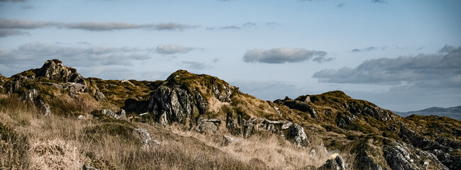 A rugged terrain features scattered rocks and dry grass, complemented by a few fluffy clouds against a bright blue sky, creating a serene atmosphere.