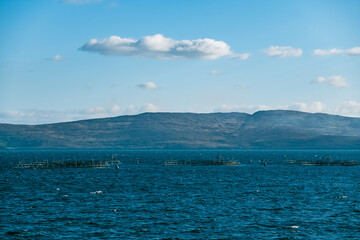 A calm blue body of water stretches out to meet a dark, mountainous horizon, with few fish farms, scattered in the foreground.