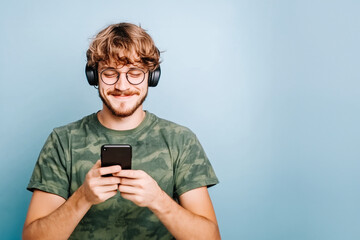 Fototapeta premium Portrait of a young man using his cell phone while wearing headphones. Isolated on blue background.