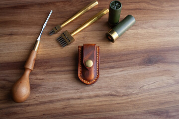 a handcrafted leather knife sheath with a brass button clasp, various leatherworking tools, all arranged on a polished wooden surface that highlights the artisanal and rugged aesthetic of the items