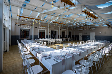 A bright, glass-walled banquet hall with tables covered with white tablecloths. Tableware ready to serve dishes. Photo taken during the day, with sunlight streaming in.