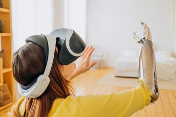Woman with bionic prosthetic hand using virtual reality glasses, immersing herself in game world of new technologies in bright modern bedroom in bright apartment