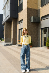 Caucasian teenager girl in a yellow shirt and blue jeans stands in front of a brick building. She is wearing headphones and has a smile on her face