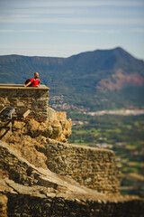 Naklejka premium A man on the edge of a medieval fortress on top of a mountain on a panoramic landscape background. Castle, Piedmont, Italy, mountains