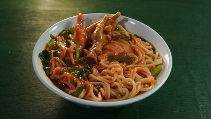 a typical Indonesian food called mie ayam ceker in a bowl on a green wooden board. Indonesian culinary, street culinary, street food, spicy noodles, noodles with chicken feet, chicken noodle