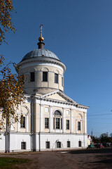 This image features an old Orthodox church with domes and a neoclassical design, including large columns and faded walls. The structure shows signs of wear, set against a bright sky and autumn trees.