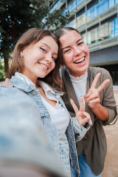 Vertical. Two happy young women smiling and posing for a selfie outdoors, capturing a fun and joyful moment together while enjoying time outside in an urban setting, showing peace sign and friendship