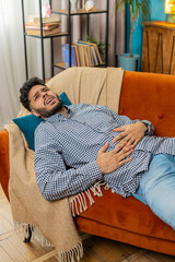 Indian young man sits on sofa feeling sudden strong abdominal stomach ache gastritis problem....