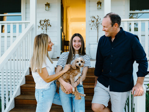 Young teenage girl carrying a dog while standing beside her parents outside the house.