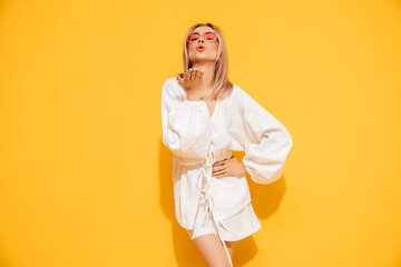 Young beautiful smiling blond female in trendy summer  white top and skirt clothes. Carefree woman posing near yellow wall in studio. Positive model. Cheerful and happy, sunglasses, gives air kiss