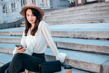 Young beautiful smiling woman in trendy summer clothes. Carefree female posing in street in sunny day. Positive model holds smartphone, looks at cellphone screen, uses mobile apps, in hat