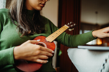  Young woman practicing ukulele guitar while reading notes on laptop