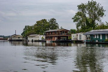 Obraz premium River Meuse with different types of houseboats moored at riverbank, floating cityscape against grey sky, leafy trees in background, cloudy day in Maastricht, South Limburg, Netherlands