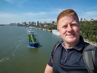 Portrait of smiling redhead caucasian man taking selfie video from bridge against large oil tanker...