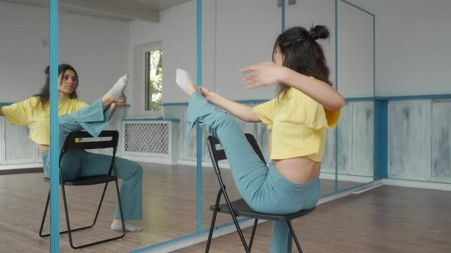 A young slender female dancer stretches her legs before starting a dance, sitting on a chair in front of a mirror in a dance studio, alternating legs. The girl is warming up and practicing dance moves