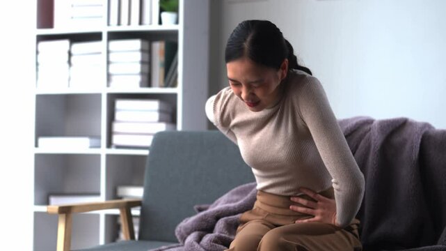 Young woman enduring discomfort from back pain while sitting on a sofa