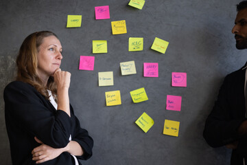 A thoughtful  businesswoman in the meeting room with her team and presenting  symbolizing success and technology while engaging with a sticky note brainstorming on a grayboard 