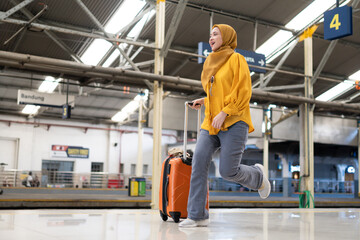 Fototapeta premium Woman in Hijab Waving at Train Station Platform. Smiling woman in a yellow hijab waves while running at a busy train station platform with her luggage. She appears excited and ready for travelling.