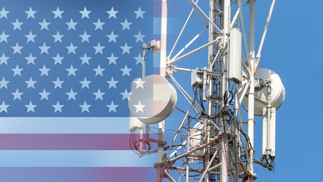 A cell phone tower is shown with the American flag in the background. The tower is surrounded by wires and is located in the middle of the image - Powered by Adobe