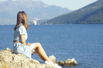 A girl in a dress sits alone on the seashore and enjoys the view of the Bay of Kotor during her vacation in Montenegro. A young woman looks at a cruise liner sailing on the sea and mountains.