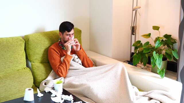 A man with a cold flu lying on the sofa at home covered with a blanket, blowing his snot with toilet paper, Sick with fever has pills and tissues on the table. seasonal virus. colorful 4k video