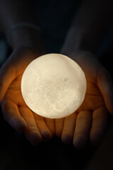 Hands holding an illuminated moon-like sphere lamp against a dark backdrop