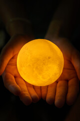 Hands holding an illuminated moon-like sphere lamp against a dark backdrop