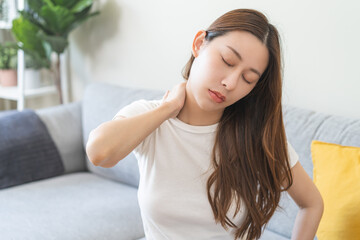 Fototapeta premium Pain body muscles stiff problem, asian young woman, girl painful with back, neck ache from work hand holding massaging rubbing shoulder hurt, sore sitting on couch at home. Health care and medicine.