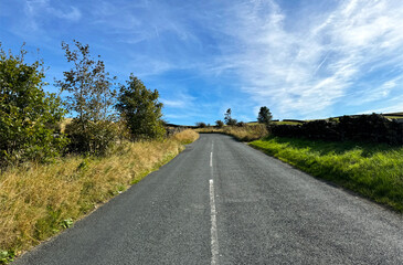 A country lane unfolds before you, bordered by lofty grasses and intermittent shrubs beneath a brilliant azure sky. The setting is tranquil and idyllic on Ellers Road, Sutton-in-Craven, UK.